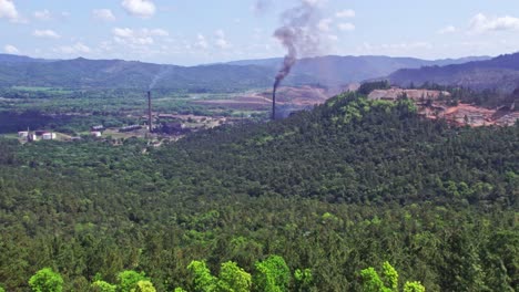 aerial forward over verdant landscape with smoking chimneys in nickel mine, loma miranda