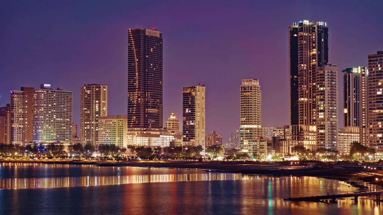A dusk-to-night timelapse view of the modern Gurney Drive waterfront, showing illuminated luxury skyscrapers and high-rise buildings reflecting on the sea