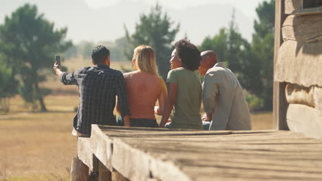 Rear-View-Of-Friends-On-Vacation-Sitting-On-Porch-Of-Countryside-Cabin-Taking-Selfie-On-Mobile-Phone