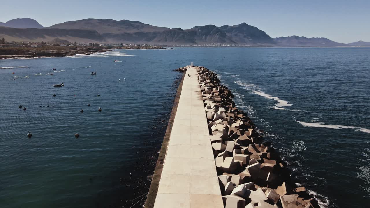 Concrete harbor dock pier in Hermanus South Africa with mountains in the distance, seagulls in flight, boats, buoys and a solid breakwater on a calm day