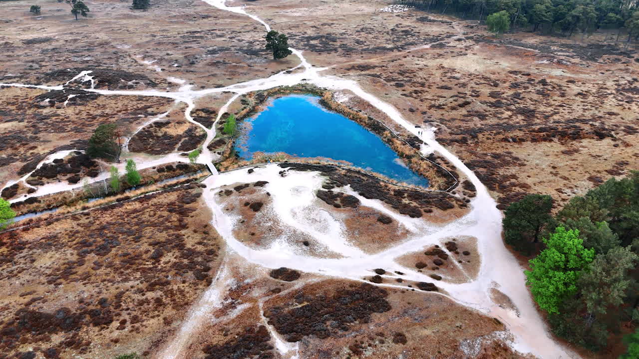 Blue sky reflecting in the little pond of triangle shape. Drone footage above the dry land approaching the lake around which two people run.
