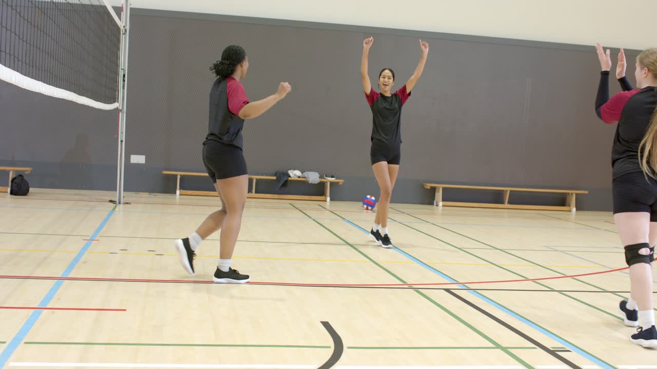 Una squadra di pallavolo femminile diversificata si tuffa vicino alla rete su un campo da palestra, con la battuta in arrivo per recuperare la palla.