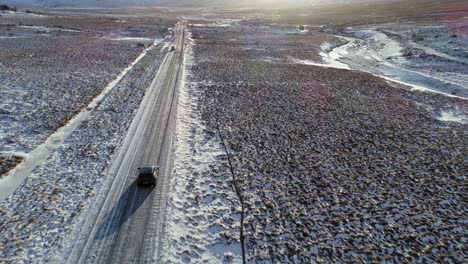 Autos,-Die-Auf-Der-Asphaltstraße-Fahren,-Umgeben-Von-Schneebedeckten-Feldern-In-Island-Mit-Malerischen-Bergen-Und-Heller-Sonne-Im-Hintergrund---Luftdrohnenaufnahme