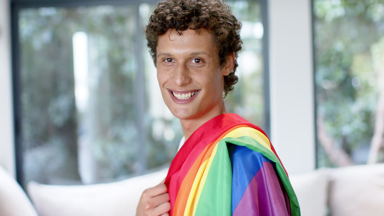 Portrait of happy biracial man holding rainbow lgbt flag at home, slow motion