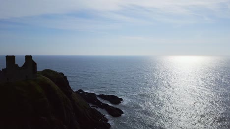 Flight-past-iconic-Dunnottar-Castle-in-Scotland,-gliding-towards-the-mesmerizing-oceanic-waves-at-sunrise