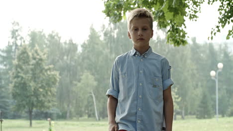 Blond-Boy-In-Blue-Shirt,-Kicks-Soccer-Ball,-Catches-It-With-His-Hands-And-Looks-At-The-Camera-In-The-Park