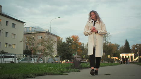 thoughtful woman in white coat holding umbrella walking slowly on paved path with residential buildings, benches, green trees, and blurred people in background