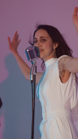 a female singer in a white gown stands in front of a vintage microphone, passionately singing with her hands outstretched. a guitarist plays in the background, and a blurred keyboardist