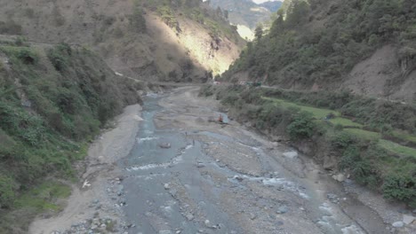 Bagger-Im-Felsigen-Flussbett,-Der-Flussgestein-Für-Die-Zementproduktion-Zieht.-Brücke,-Die-Sich-über-Das-Wasser-Im-Tal-Erstreckt-Und-Gemeinden-In-Kabayan-Benguet,-Philippinen,-Verbindet.-Rückwärtsgang-Zeigt-Aufsteigende-Luftaufnahme