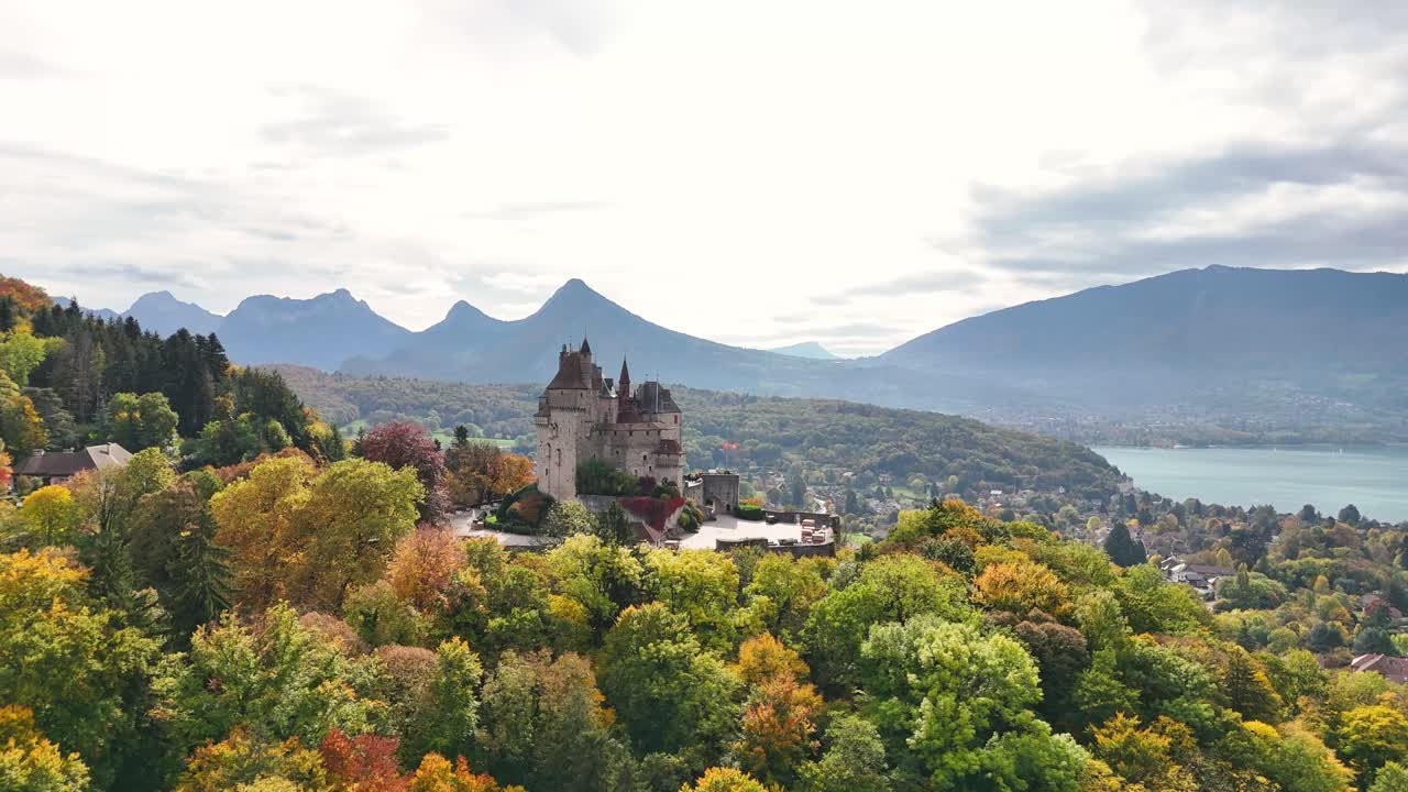 Burg Menthon-Saint-Bernard in France stands above vibrant autumn trees, framed by mountains and a calm lake. The medieval castle dominates the hillside, offering a view of the surrounding landscape