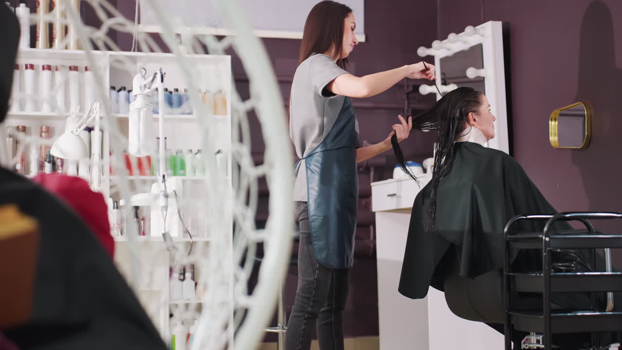 스타일리스트가 현대적인 보라색 살롱에서 백인 고객의 긴 머리를 다듬고 있어요. 앞치마를 입은 스타일리스트는 거울 근처에서 가위와 빗을 사용하고 있으며, 병과 제품 선반이 보입니다. 집중된 정밀함과 친근함이 느껴져요.