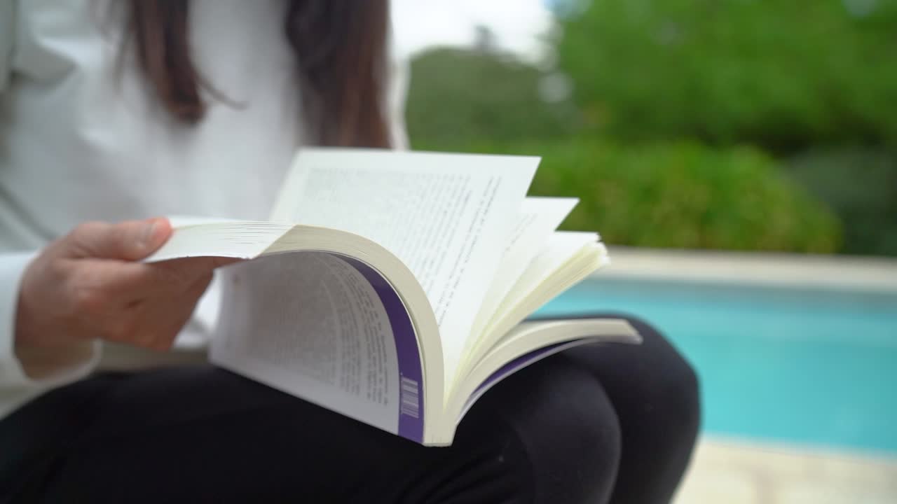 vista recortada de una mujer girando las páginas del libro sentada a orillas de la piscina