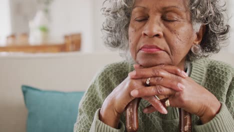 Portrait-of-tired-senior-african-american-woman-sitting-on-sofa,-leaning-on-walking-cane