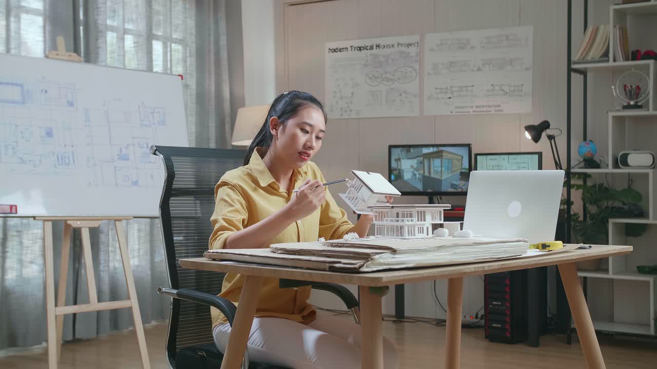 Asian Woman Engineer With A Laptop Thinking While Holding Paper Model Of House At The Office