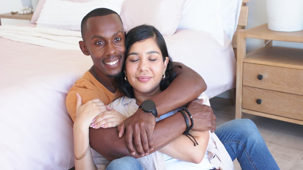 Casal diverso sentado no chão perto da cama, homem sorrindo, apertando o abraço e mulher com os olhos fechados