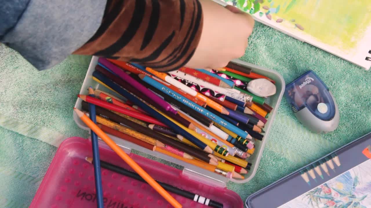 Lifestyle shot of a female adult's hand going through her pencil box, searching for green colored pencil crayons in outdoor environment at daytime.