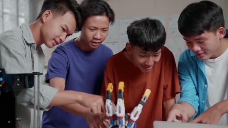 grupo de jóvenes chicos asiáticos con una computadora portátil e impresión 3d sonriendo y cruzando los brazos a la cámara mientras discuten sobre una mano cyborg en casa