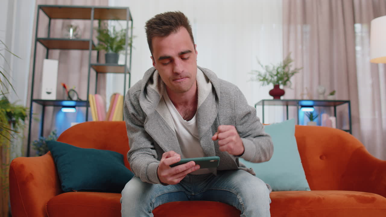 hombre joven divertido preocupado niño jugando shooter videojuegos en línea celebrar la victoria en el teléfono inteligente en casa