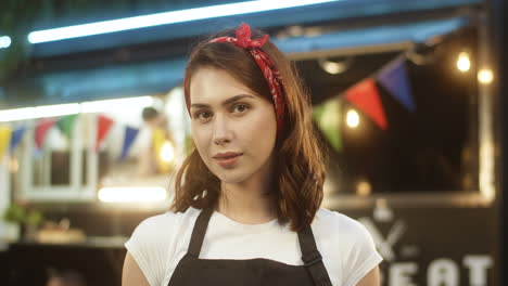 Close-Up-Of-Beautiful-Young-Woman-In-Apron-Smiling-Happily-To-Camera-Outdoors-With-Little-Food-Truck-Bar-On-Background