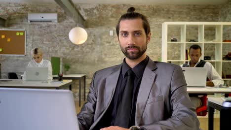 confident businesswoman with beard is sitting at laptop in office, watching at camera, colleagues are networking with technologies, work concept, communication concept