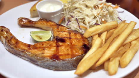 a plateful of flavourful food served in a restaurant, a combinaion of grilled salmon steak, french fries, and coleslaw salad