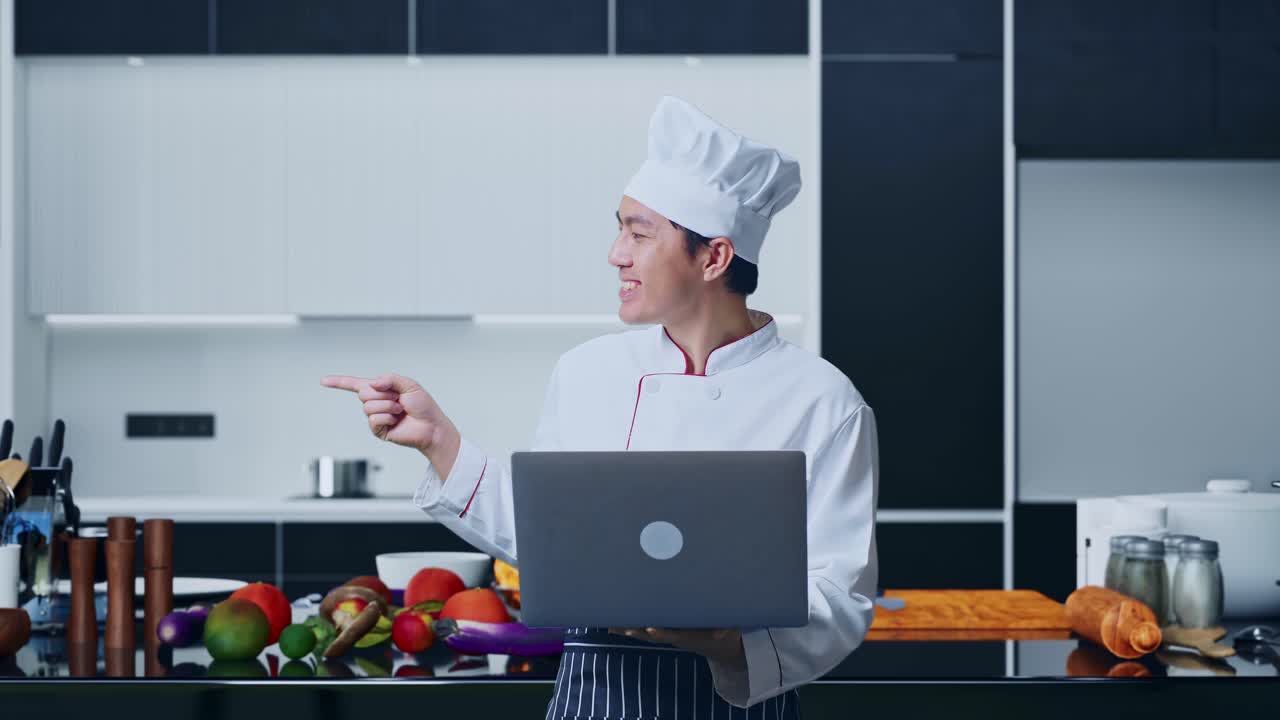 cocinero asiático usando una computadora portátil y apuntando a un lado mientras está de pie en la cocina de casa