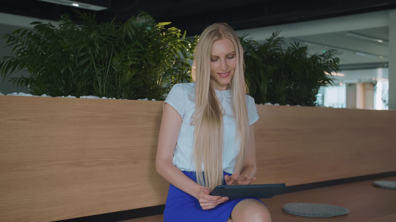 Relaxing woman with tablet in office. Young elegant woman sitting barefoot on wooden tribune in modern office using tablet and taking rest