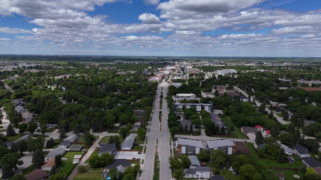 Ripresa aerea con drone dall'alto di una zona residenziale a Steinbach, Manitoba, che sorvola strade con case, alberi, erba verde, auto e cieli estivi parzialmente nuvolosi sulla piccola città canadese.