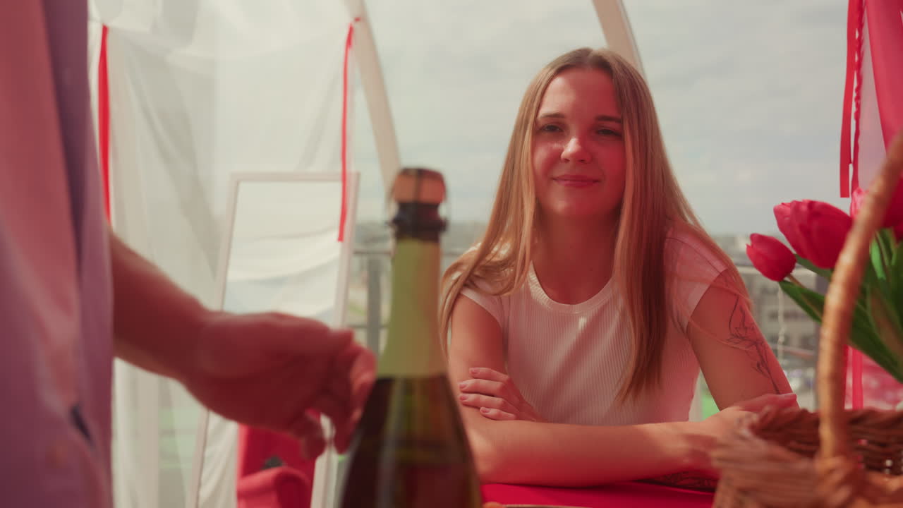 Mujer sonriente en una cita nocturna, su pareja abriendo una botella de champán junto a un ramo de tulipanes, una cesta de mimbre y ropa de cama roja, terraza iluminada por el sol con vistas al mar, tonos dorados cálidos, mirada sincera y afectuosa