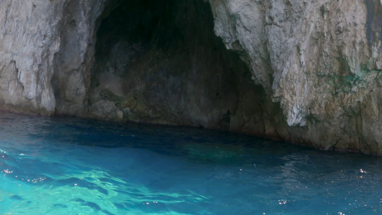 POV de um barco se aproximando da entrada de uma caverna azul no mar, litoral rochoso com água turquesa cristalina, natureza mediterrânea