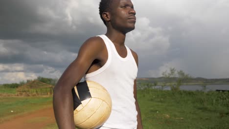 Eine-Aufnahme-In-Zeitlupe,-Die-Einen-Jungen-Afrikanischen-Mann-Umkreist,-Der-Einen-Fußball-Hält,-Während-Er-Steht-Und-Mit-Gewitterwolken-In-Der-Ferne-Zum-Horizont-Schaut