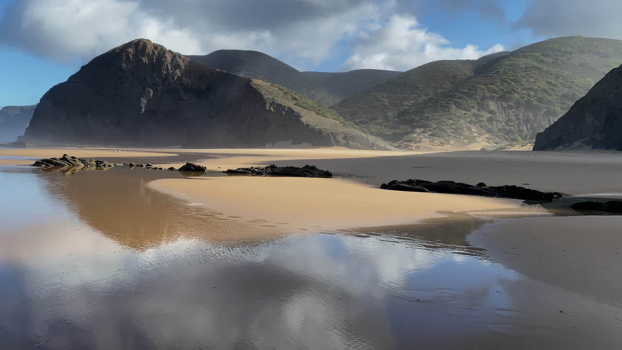 Le montagne di Sagres si riflettono sulla marea calma, creando un perfetto specchio di luce e silenzio. La sabbia dorata e le scogliere blu si incontrano sotto una luce soffusa, in un tranquillo mattino sulle spiagge deserte di Sagres.
