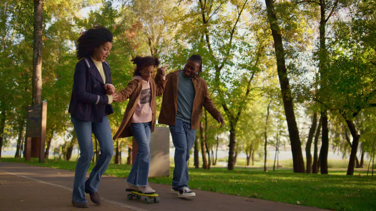 Foreldre lærer jente å kjøre longboard i parken. Støttende familie oppmuntrer barnet.