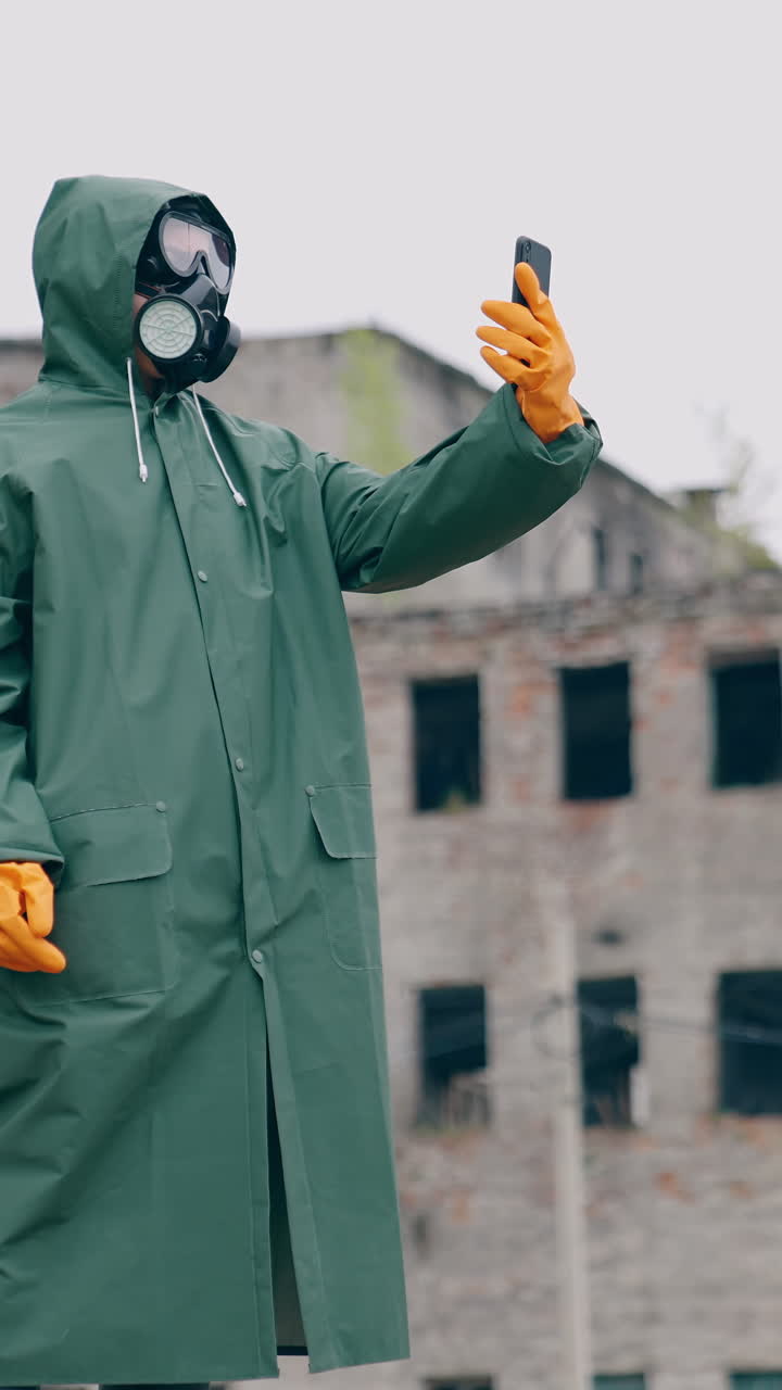Human in a protective suit and gas mask with a phone. Unrecognizable person in safety uniform, gloves and respirator doing selfie in the destroyed place during radiation. Vertical video