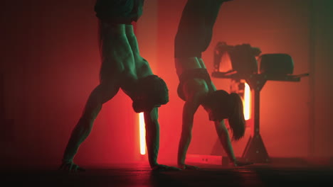a man and a woman do a handstand in the gym together in slow motion. a strong man and a woman in a handstand walk across the gym floor in colored oil lamps