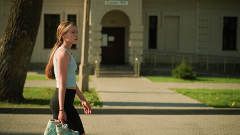 side view of young lady walking by building holding roller skates, wearing casual athletic wear under bright sunlight, background includes a tree and lush greenery