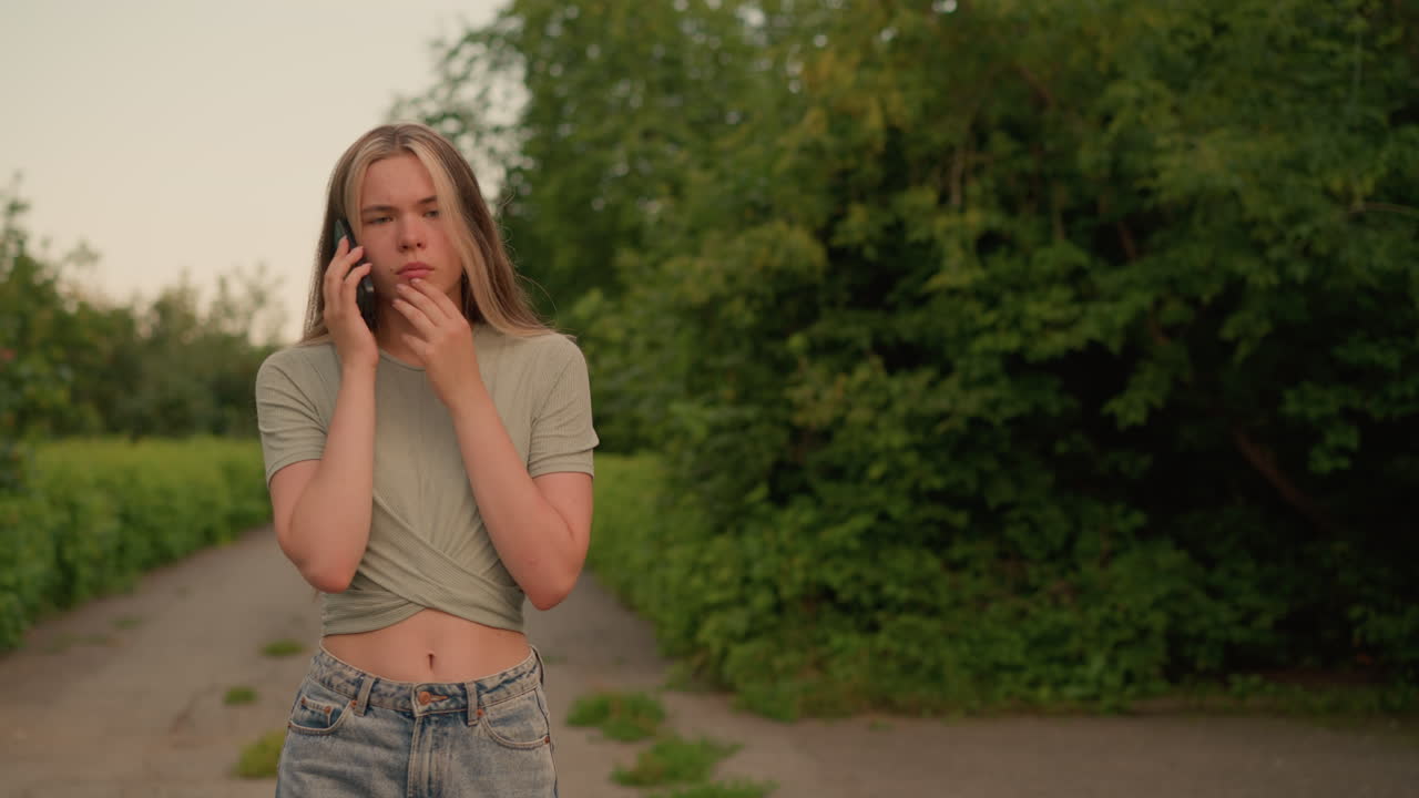 Young woman in casual jeans and grey crop top walking on path surrounded by lush greenery and trees, engaged in thoughtful phone conversation with one hand touching her jaw