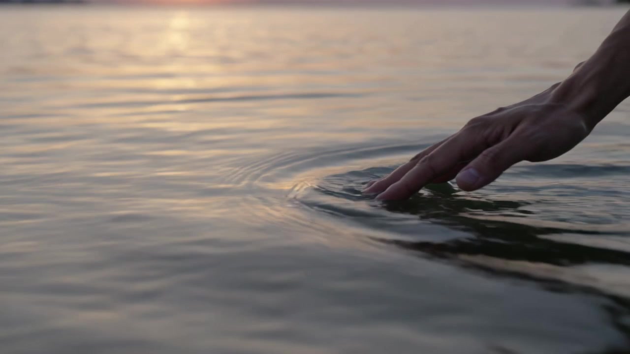 Mano che entra da destra e tocca l'acqua, creando increspature circolari che riflettono il bagliore del sole sul lago al tramonto, con spazio per il testo.