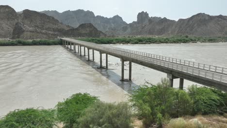 aerial drone shot over a road bridge over a river flowing with mountains in the background in hingol, balochistan, pakistan on a cloudy day