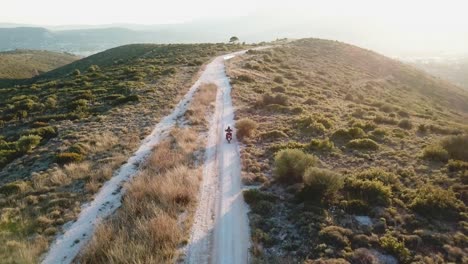 Aerial-shot-of-following-a-motorcyclist-while-he-drives-on-a-gravel-road-during-golden-hour-on-a-mountain-next-to-Athens,-Greece