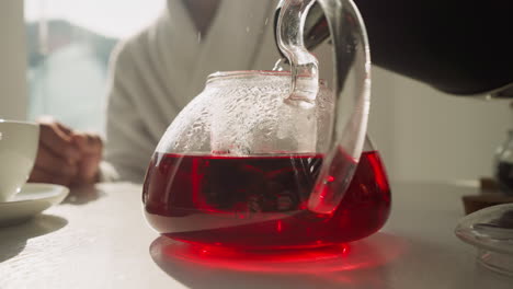 brewing hibiscus tea in teapot on table. careful preparation of nutritious herbal tea using hot flow from electric pot in morning. family ritual