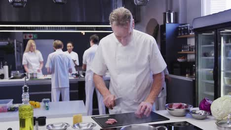 Chef-Masculino-Caucásico-Profesional-Vistiendo-Chefs-Blancos-En-La-Cocina-De-Un-Restaurante-Cortando-Carne-En-Un-Cutt