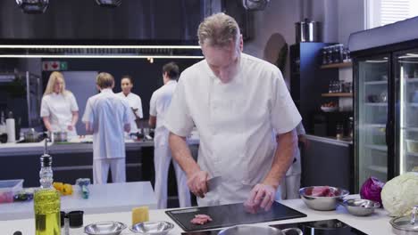 professional caucasian male chef wearing chefs whites in a restaurant kitchen slicing meat on a cutt