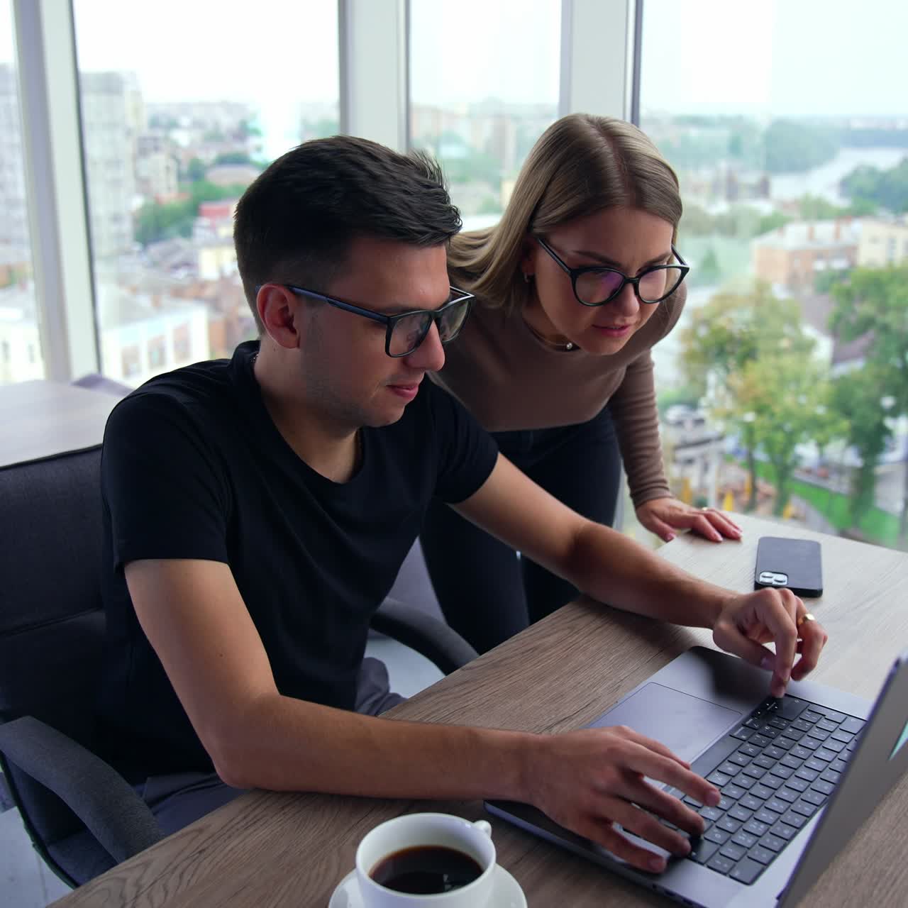 A pair of colleagues looking attentively at the screen of laptop. Colleagues are busy with doing some work on computer. Office indoor backdrop with cityscape at windows