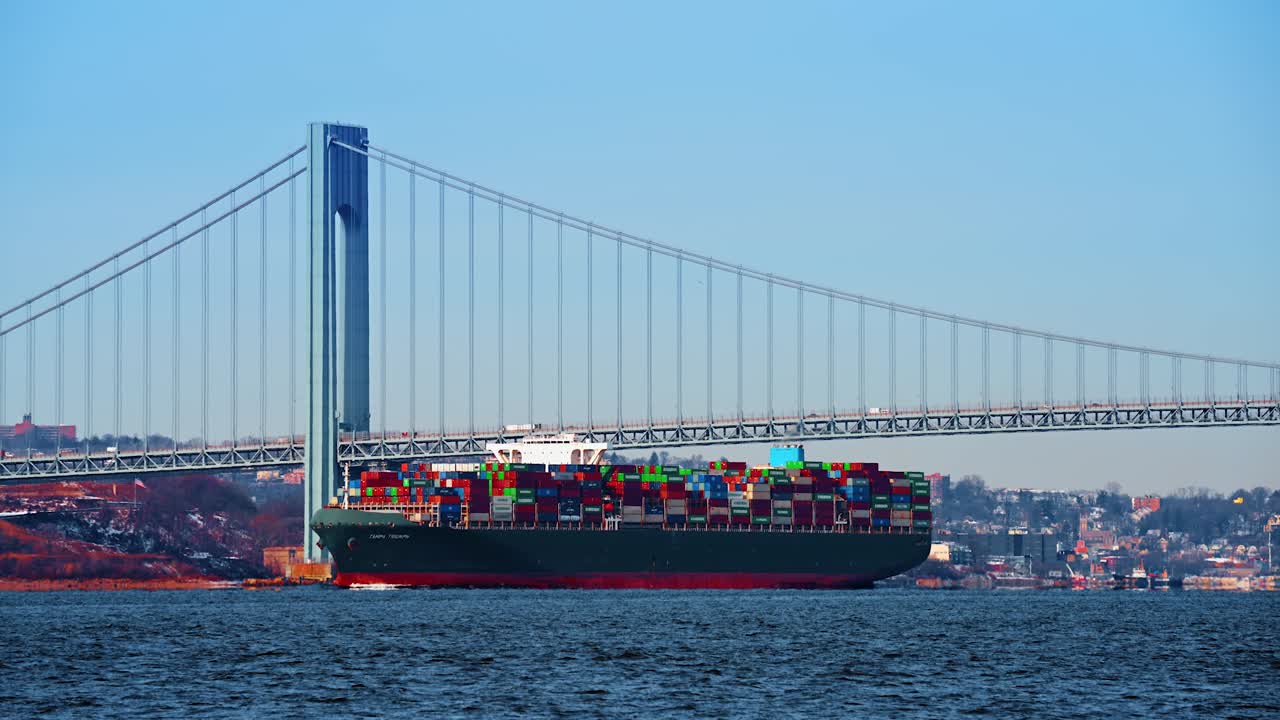 New York, USA, 5 October 2025: Cargo ship navigating under a suspension bridge. Wide shot of a green container carrier passing through a strait under a large steel bridge