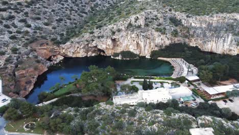 Vista-De-Pájaro-De-ángulo-Alto-Del-Lago-Sumergido-De-Vouliagmeni-En-Atenas,-Grecia-Con-Impresionantes-Acantilados-Circundantes-Y-Una-Carretera-Sobre-El-Lago-|-4k