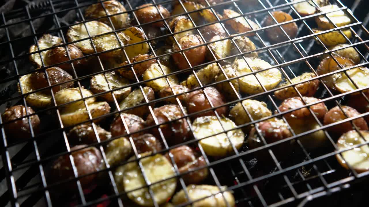 아래에서 빛나는 붉은 불씨가 있는 뜨거운 그릴에 있는 철망 바구니에 갈색 허브 새 감자를 뒤집습니다.