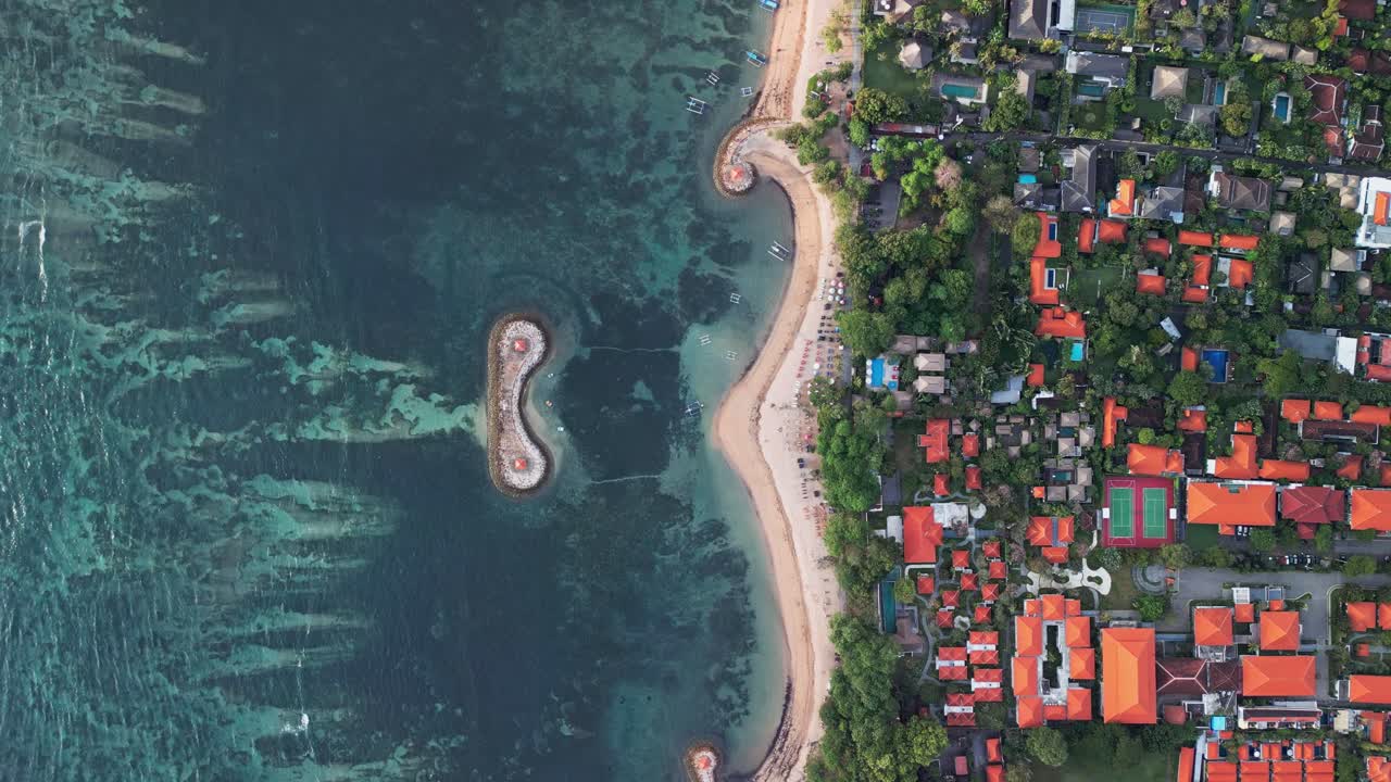 Aerial drone footage of Sanur Bali coastline with sandy shore, coral reef barriers, turquoise water, palm trees, coastal village, lush greenery, sunrise light, and peaceful tropical seascape