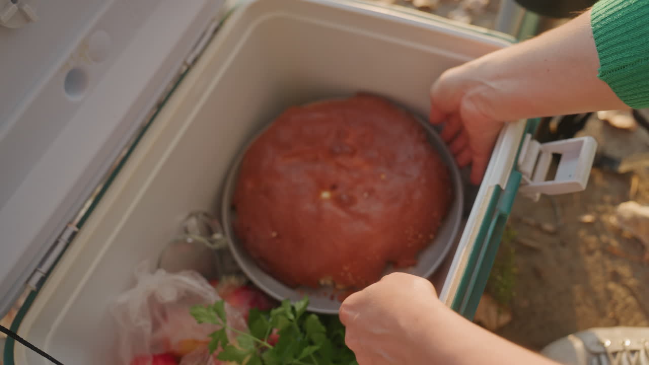 Camping cooler opens to reveal homemade cake. Camper in green sweater unlatches insulated cooler beside thermos, lifts round golden cake on metal pan onto cooler lid, autumn leaves underfoot, warm