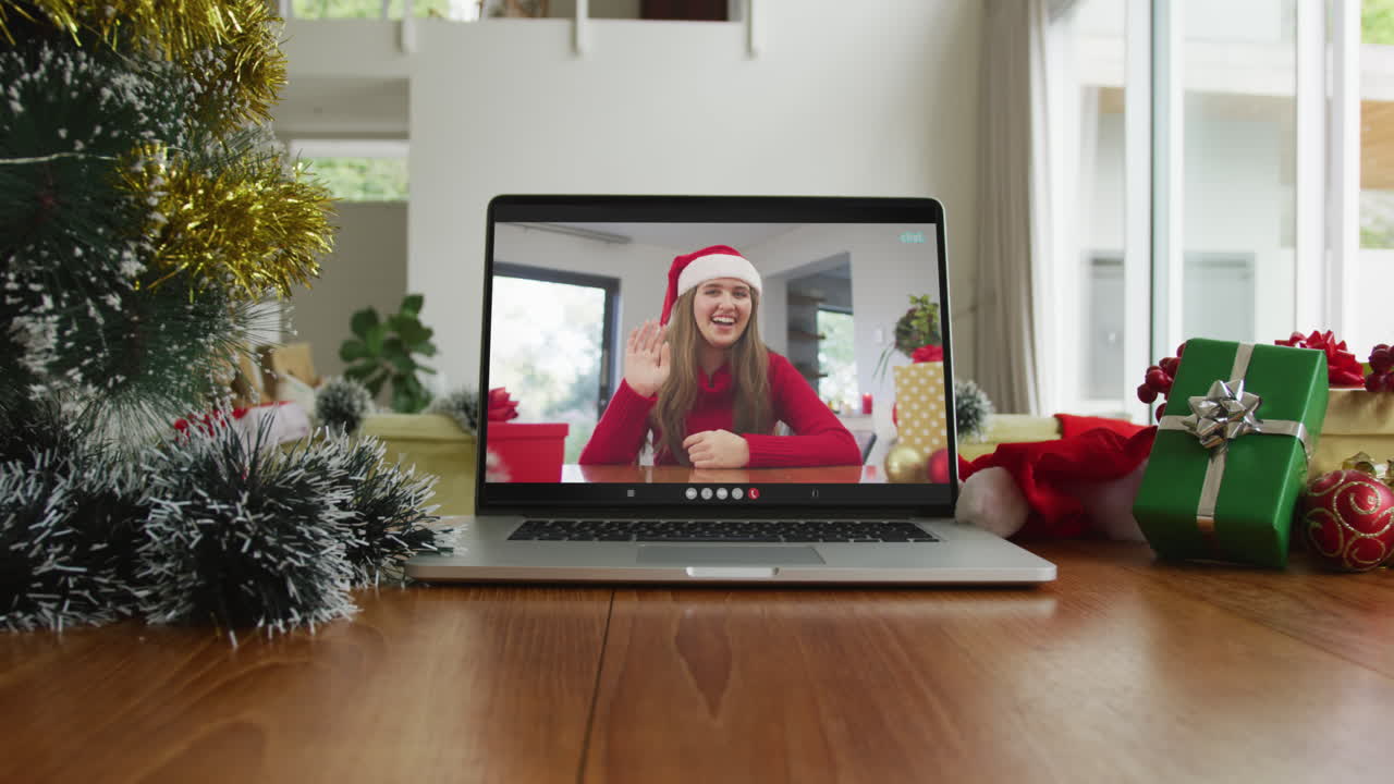 mujer caucásica sonriente con sombrero de papá noel y saludando en la videollamada de navidad en la computadora portátil